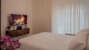 Ein Schlafzimmer mit einem Bett, einem an einer Holdekor-Kommode montierten Fernseher, einer Kristallleuchtre und transparenten Vorhängen.