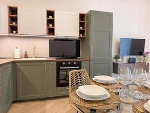 A compact kitchen with built-in cabinets, a stove, and a dining area with wicker chairs and plates. - Velora Roma Apartment [Vatican Area] Elegant & Intimate Retreat in Rome (Roma)