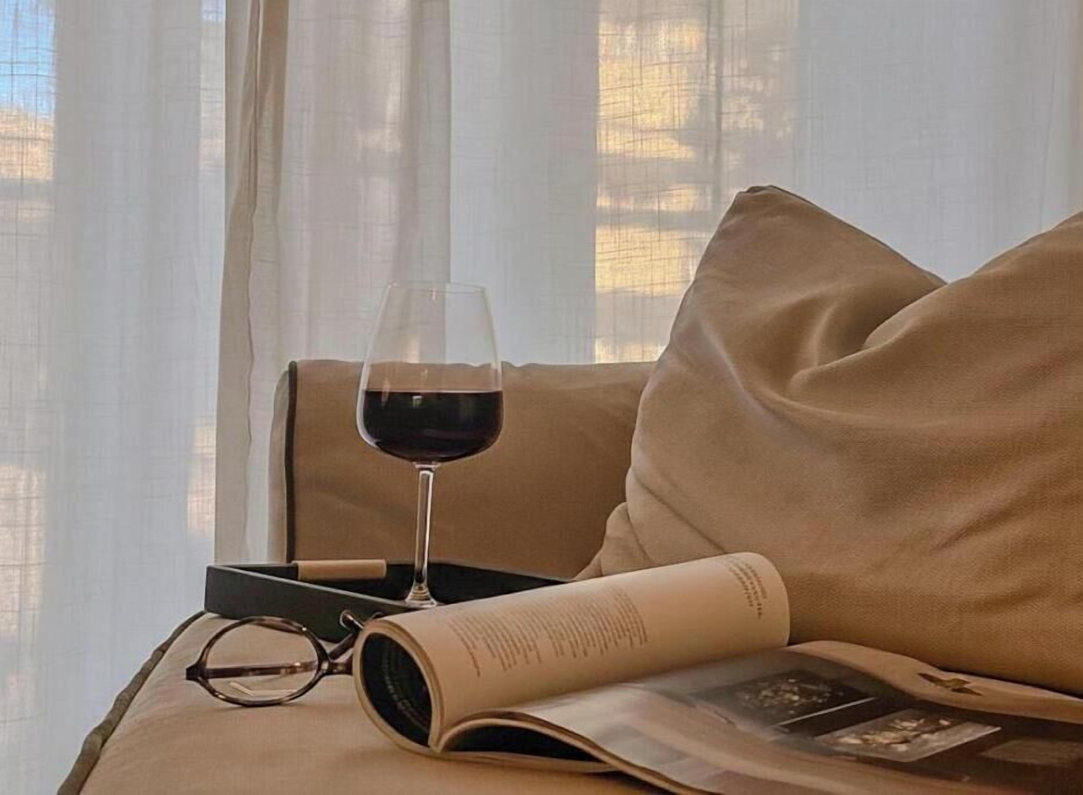 A room with a window, a glass of red wine, a book, and a pair of glasses on a beige sofa.