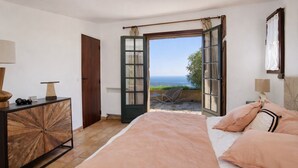 Un dormitorio con cama, cómoda de madera, puerta abierta que da a un balcón y vista al mar.