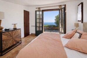 Une chambre à coucher avec un lit, une commode en bois, une porte ouverte donnant sur un balcon et une vue sur la mer.