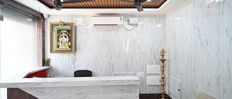 Un área de recepción con mostrador de mármol, ventiladores de techo, unidad de aire acondicionado, estatua religiosa enmarcadas y una lámpara tradicional.