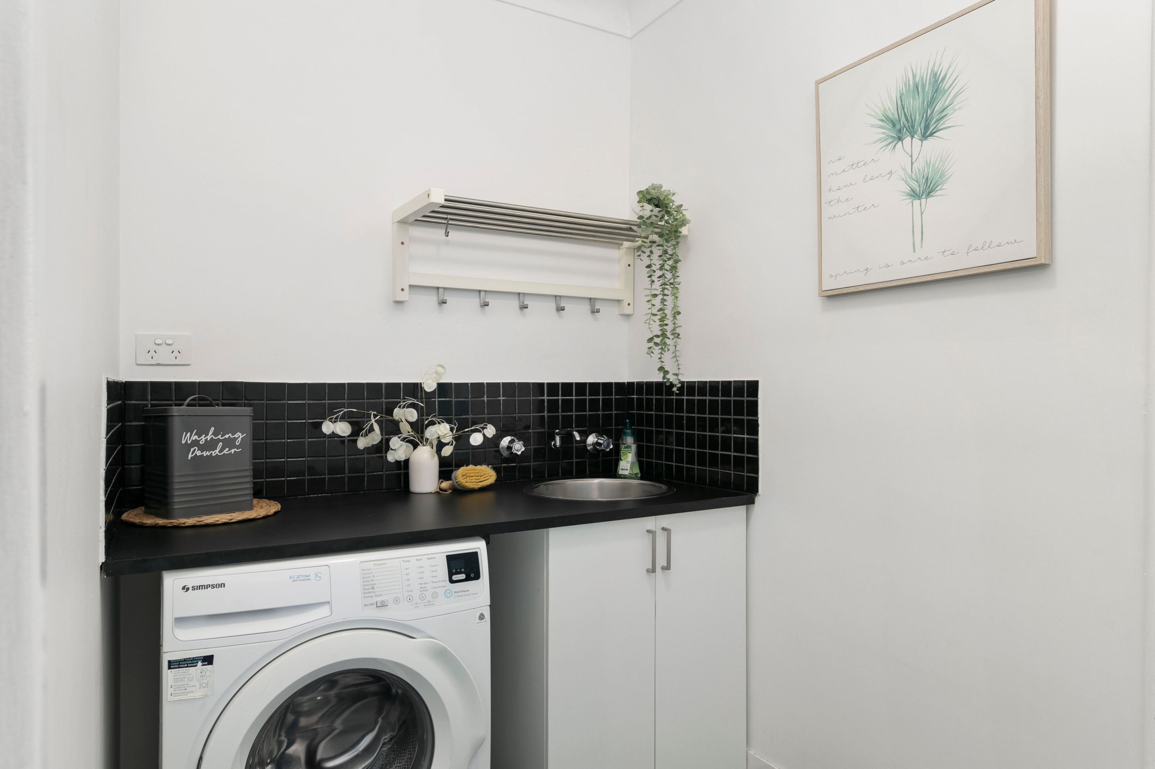 A compact kitchen with a washing machine, a hanging plant, and a framed quote on the wall.