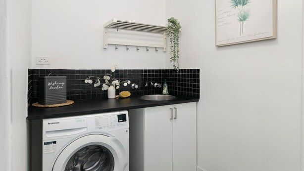A compact kitchen with a washing machine, a hanging plant, and a framed quote on the wall. - Vino Cottage (Cessnock)