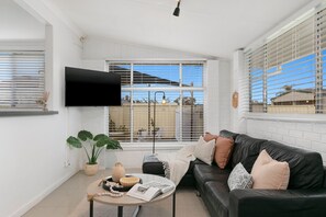 A modern living room with a black leather sofa, a round coffee table, and a flat-screen TV mounted on the wall. - Vino Cottage (Cessnock)
