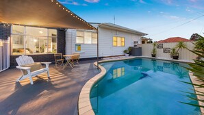 A residential swimming pool with a covered patio area, outdoor furniture, and a clear blue sky. - Vino Cottage (Cessnock)