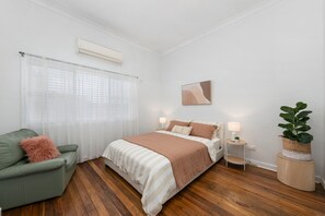 Ein Schlafzimmer mit einem Bett, einem grünen Sessel, einem Nachttisch mit Lampe, einer Topfpflanze und einem Fenster mit transparenten Vorhängen.