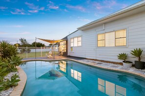 A swimming pool with a clear blue water surface, surrounded by a white house with large windows and a covered patio area. - Vino Cottage (Cessnock)