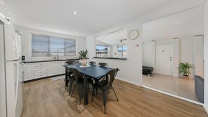 A modern kitchen with white cabinetry, a dining table with chairs, and a large window with blinds. - Vino Cottage (Cessnock)