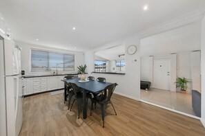 A modern kitchen with white cabinetry, a dining table with chairs, and a large window with blinds. - Vino Cottage (Cessnock)