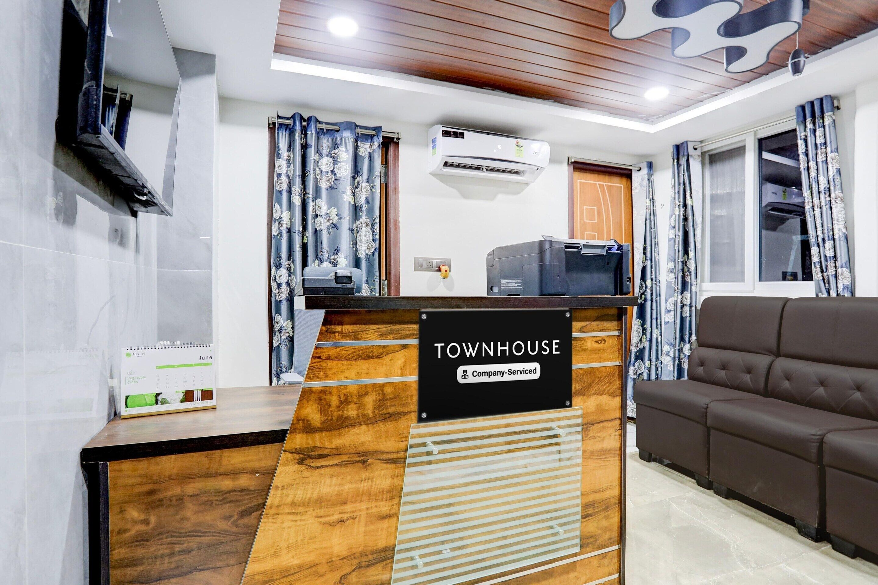 A reception desk with a sign reading 'TOWNHOUSE Company-Serviced,' a flat-screen TV, an air conditioning unit, a calendar, and a sofa in the background.