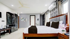 Ein modernes Hotelzimmer mit einem großen Bett, einem Nachttisch aus Holz, einem Schreibtisch mit Stuhl, einem Deckenventilator und einer Klimaanlage.