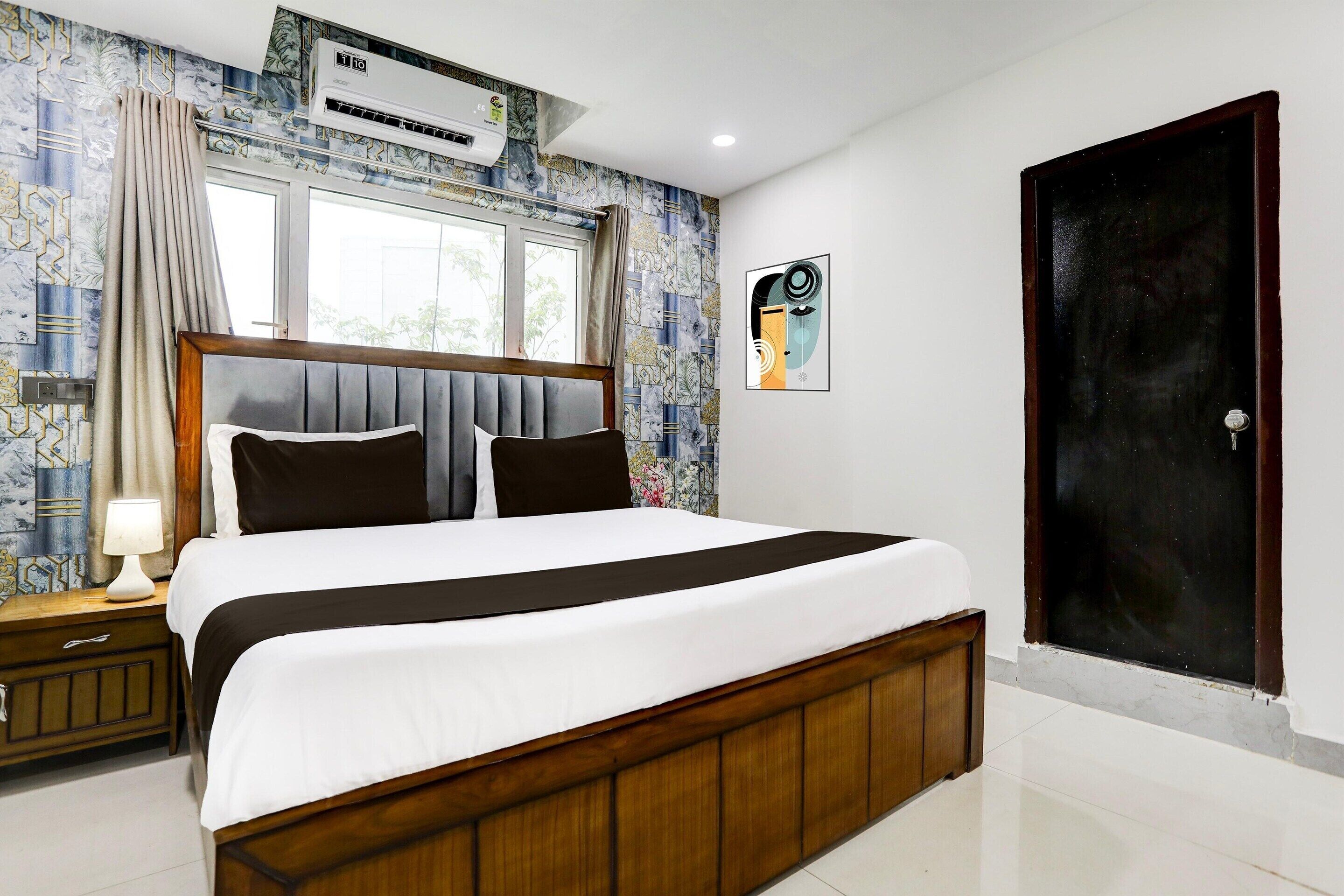 Habitación de hotel con una cama grande, una mesita de noche con lámpara, una ventana con cortinas, un equipo de aire acondicionado y una puerta.