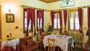 Un coin repas avec une table dressée pour deux personnes, entourée de chaises, des rideaux rouges aux fenêtres et un lustre au-dessus.