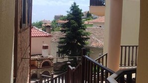 Une vue à travers une fenêtre en arc d’un bâtiment dont un escalier mène à un balcon, d’où l’on aperçoit un arbre et des bâtiments aux toits recouverts de tuiles rouges.