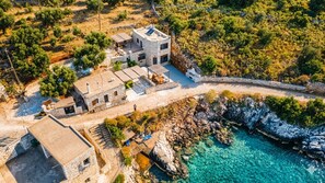 Vue aérienne d’une propriété côtière comprenant une maison en pierre, un bâtiment séparé et une côte rocheuse bordée d’une eau d’un bleu limpide.