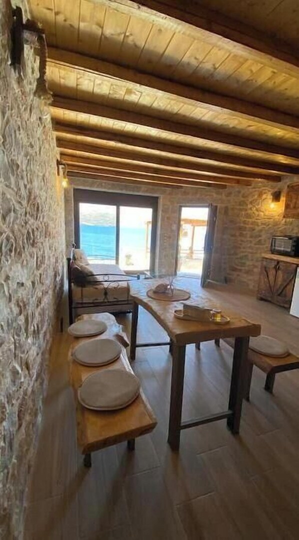 Un intérieur rustique avec un mur en pierre, des poutres en bois, une table avec des chaises et une vue sur la mer par de grandes fenêtres.