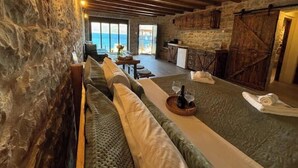 A cozy room with a stone wall, wooden ceiling beams, a sofa, a dining table with a glass top, and a view of the sea through large windows.