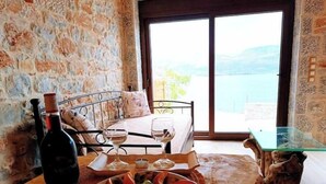 A room with a stone wall, a bed with a view, a table with a plate of olives and grapes, and a bottle of wine.