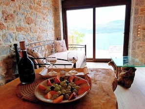 Habitació amb paret de pedra, llit amb vistes, taula amb un plat d'olives i raïm, i una ampolla de vi.