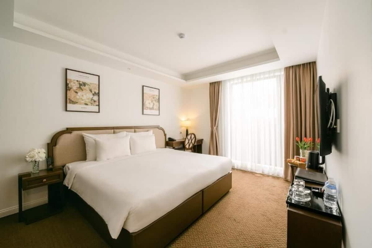 A hotel room with a large bed, a bedside table with a lamp, a TV mounted on the wall, and a window with curtains.