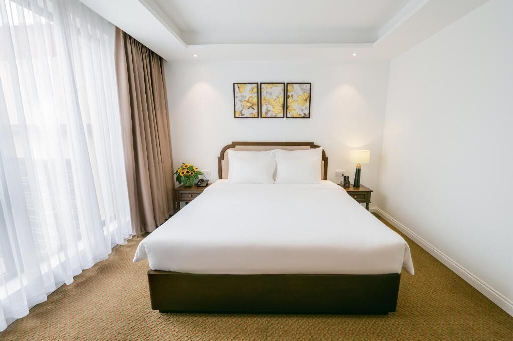 Habitación de hotel con una cama grande, una mesita de noche con una lámpara, un jarrón con flores y una ventana con cortinas de encaje.