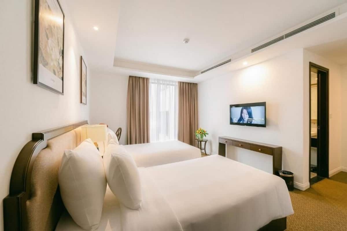 A hotel room with two beds, a television, a window with curtains, and a small table with a vase.