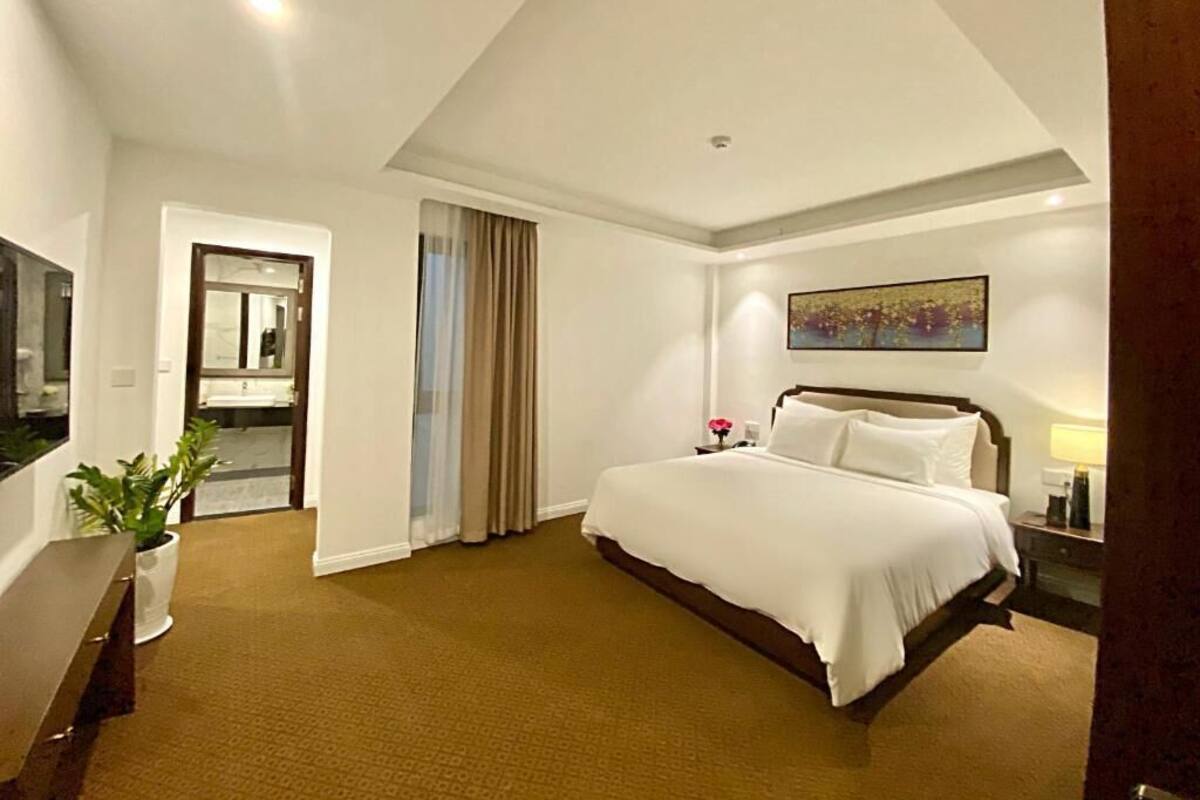 A hotel room with a bed, a desk, a chair, a potted plant, a painting, and a door leading to a bathroom.