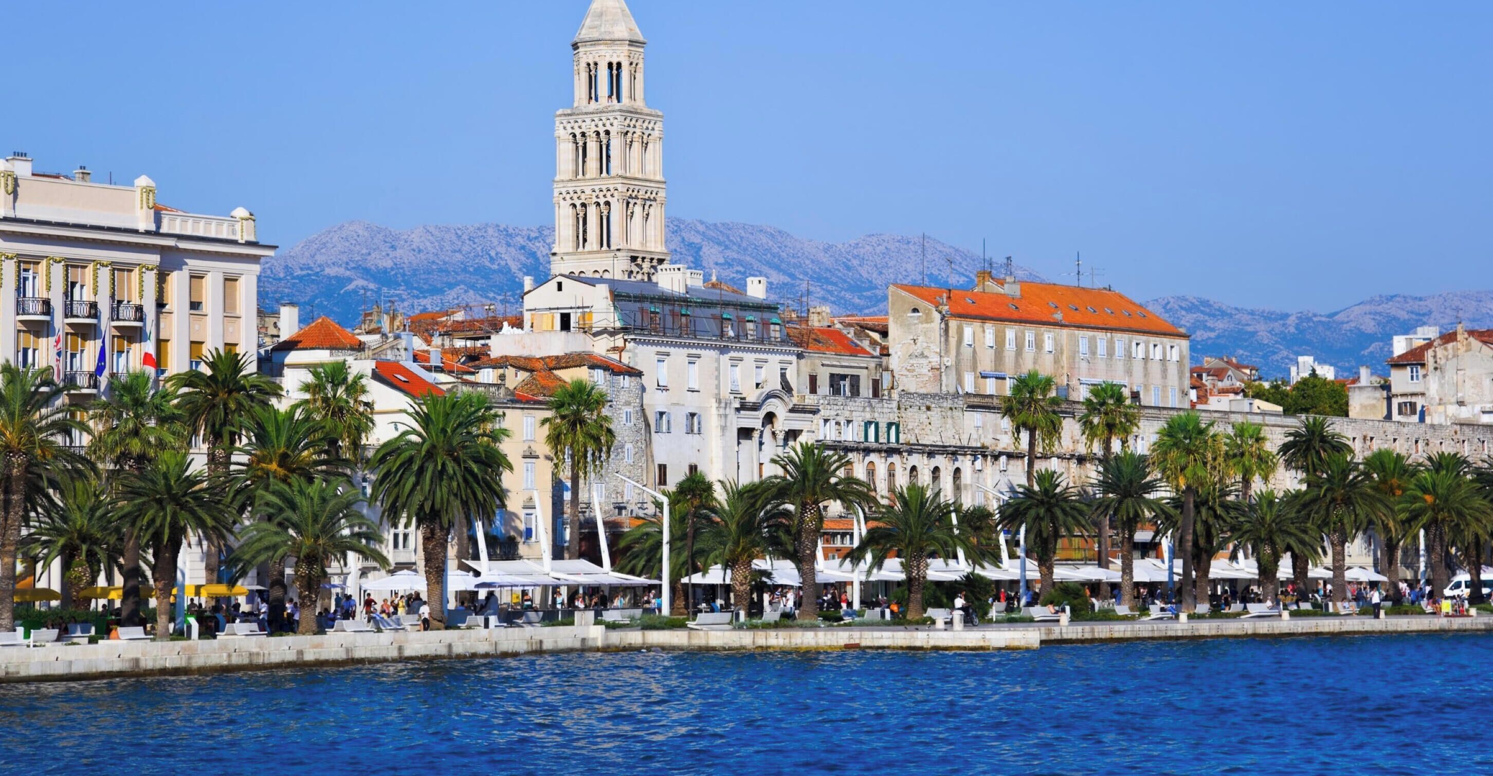 A coastal cityscape with a prominent tower, historic buildings, and a waterfront promenade with palm trees.