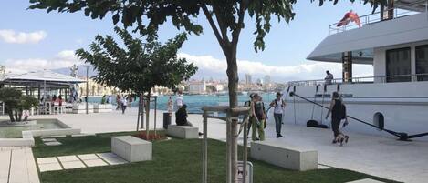 A waterfront promenade with seating areas, a large tree, and a white boat docked nearby.