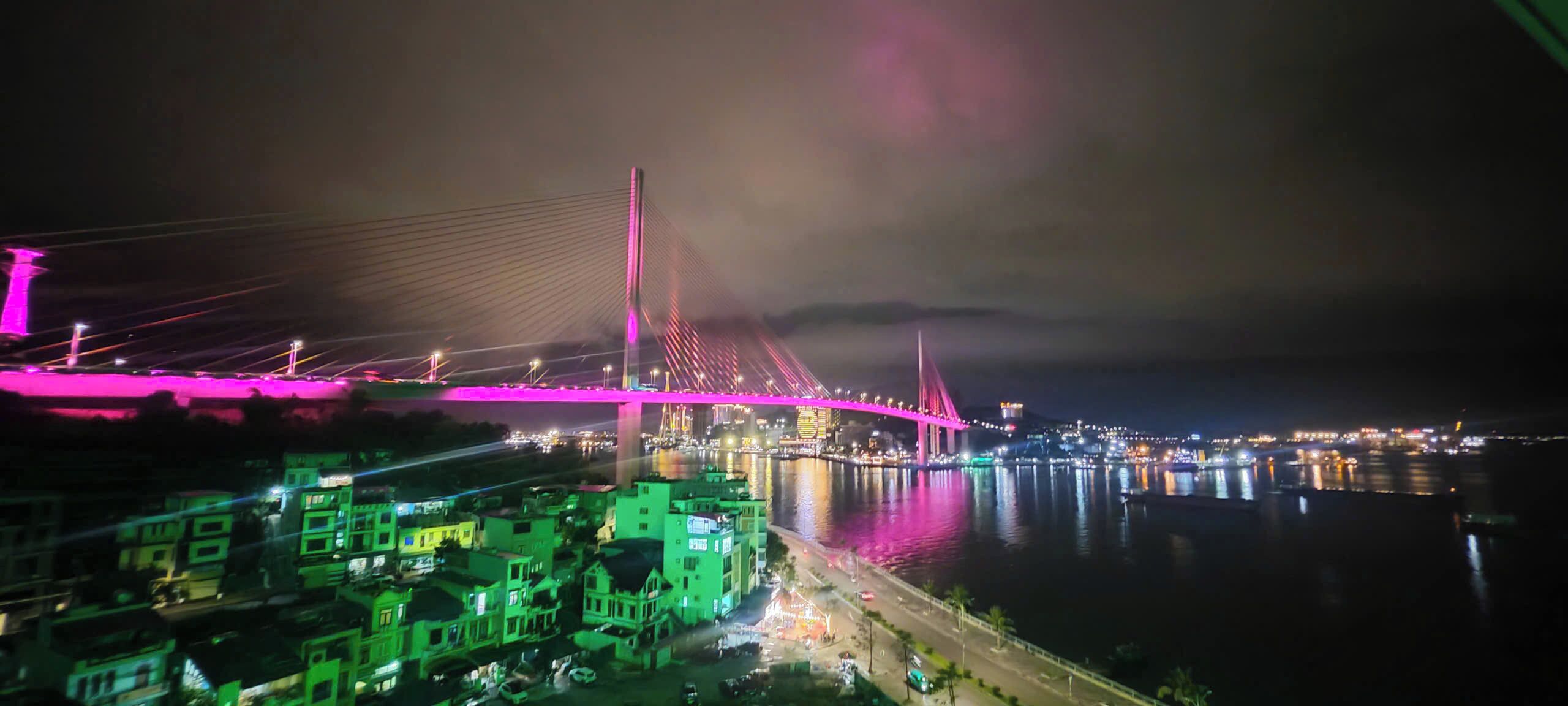 A nighttime view of a bridge with illuminated cables and a river reflecting lights.