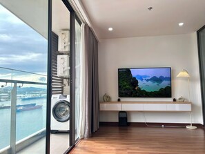 A room with a large window overlooking a body of water, a washing machine, a TV on a wall, and a small table with a lamp. - Jade Leaves Homestay HaLong -Seaview (Hong Gai)