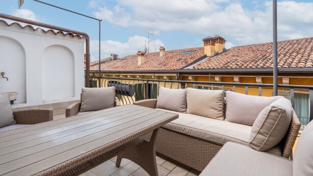 A patio with wicker furniture, a table, and a view of buildings with red-tiled roofs. - Elegant residence overlooking Porta Leoni (Verona)