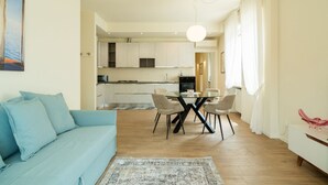 A modern living room with a sofa, a dining table with chairs, a kitchen in the background, and a large window with curtains. - Elegant residence overlooking Porta Leoni (Verona)