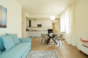 Ein modernes Wohnzimmer mit einer Couch, einem Esstisch mit Stühlen, einer Küche im Hintergrund und einem großen Fenster mit Vorhängen.