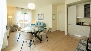 A modern living room with a glass dining table, light-colored sofas, a large window with curtains, and a kitchen area with cabinets. - Elegant residence overlooking Porta Leoni (Verona)