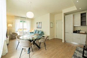 Ein modernes Wohnzimmer mit einem Glastisch, hellen Sofas, einem großen Fenster mit Vorhängen und einer Küchenzeile mit Schränken.