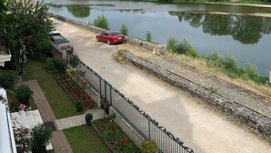 Vista al río con un sendero pavimentado, un coche rojo estacionado junto al río y un edificio con pared de ladrillo y fachada blanca.