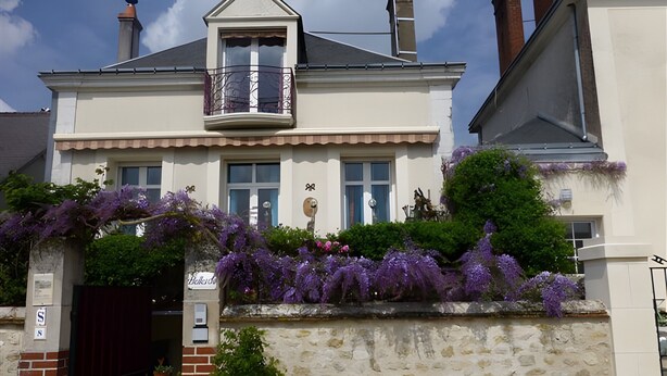 Un edificio de dos pisos con un balcón, un muro de piedra y enredaderas moradas con flores.