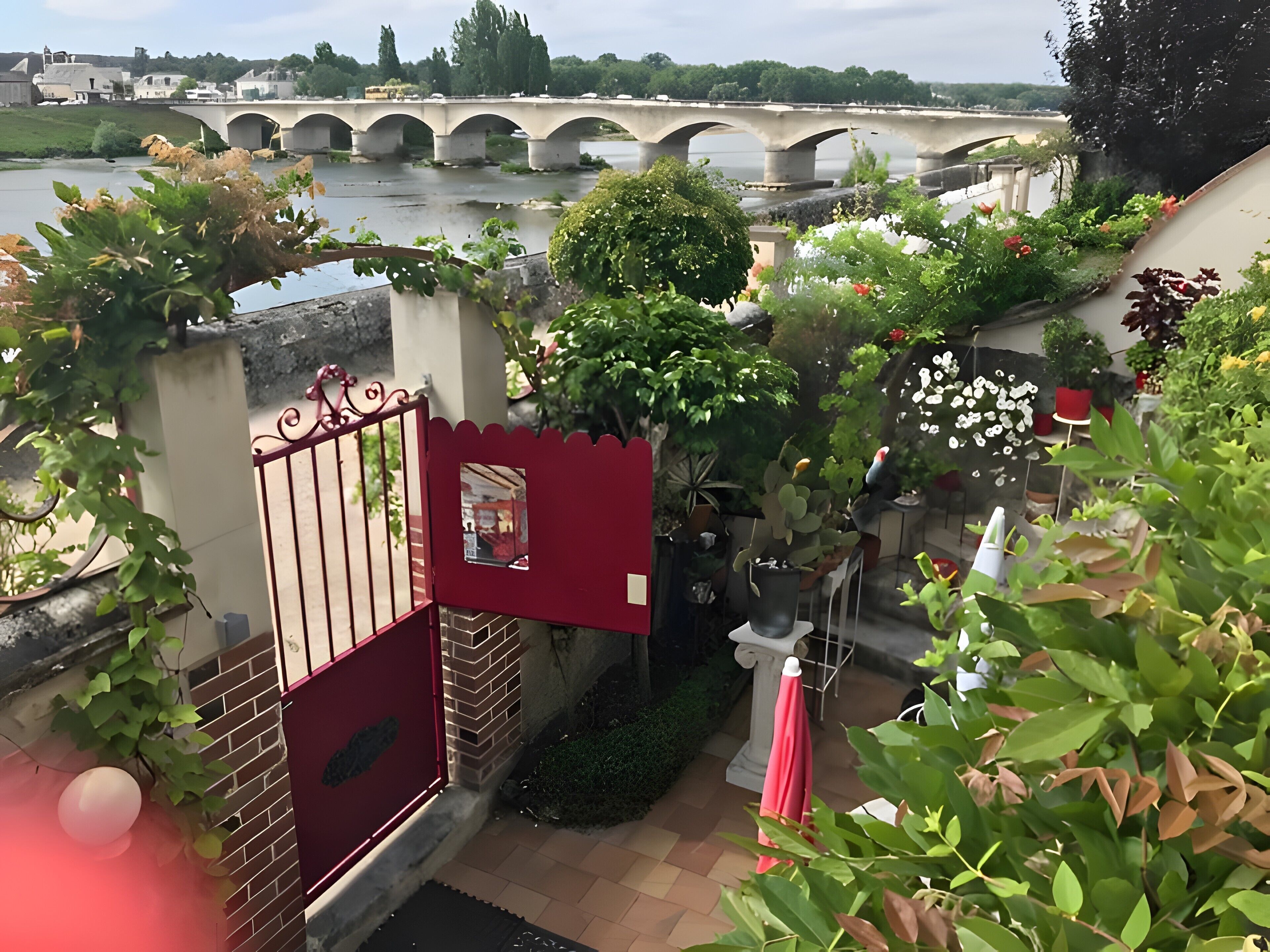 Un jardin avec une porte rouge, des plantes en pot et une vue sur un pont qui enjambe une rivière.