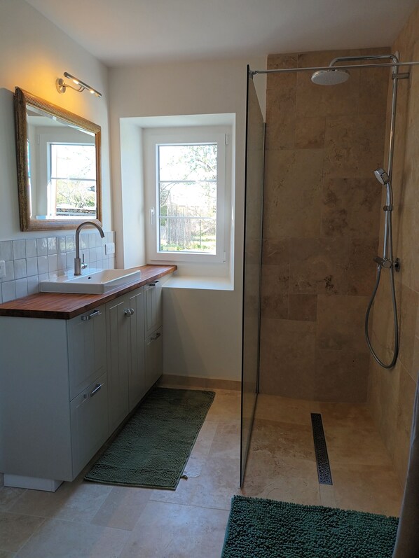Une salle de bain moderne équipée d’une douche, d’un lavabo avec un miroir et d’une fenêtre laissant entrer la lumière naturelle.