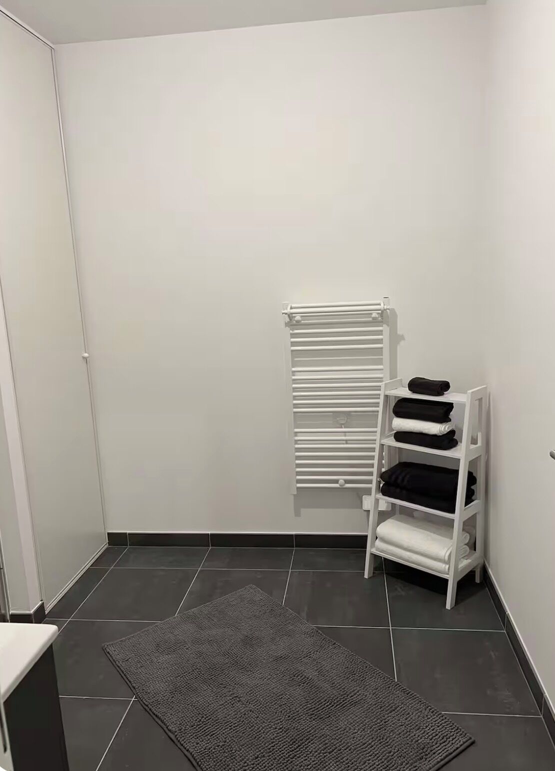 Une petite pièce propre, avec un radiateur blanc, un tapis gris et une étagère blanche où sont pliées des serviettes.