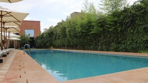 Una piscina con lettini e ombrelloni, immersa nel verde e affiancata da un edificio moderno.