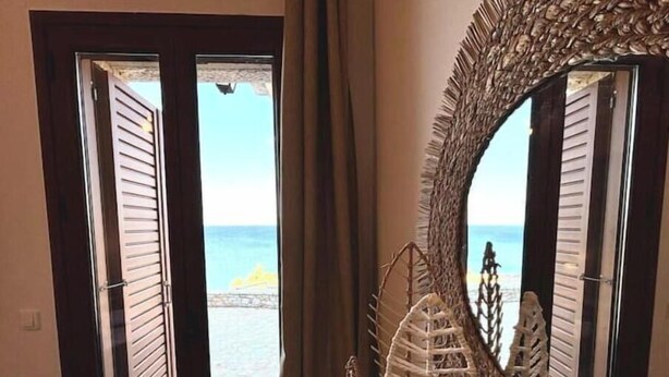 Quarto com vista para o mar através de uma porta com venezianas horizontais, um espelho grande com moldura texturizada e um objeto decorativo na parede.