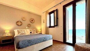 Quarto com cama, duas mesas de cabeceira, relógio na parede e vista para o mar através de uma porta deslizante.