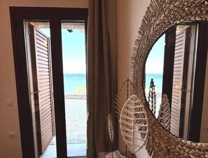 A room with a view of the sea through a door with horizontal blinds, a large mirror with a textured frame, and a decorative wall piece. - Magic estate on the wave 2 (Kotronas)