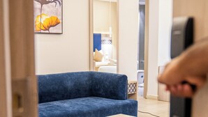A modern hotel room with a blue sofa, a small wooden coffee table, and a mirror reflecting a bathroom with a towel rack. - Al Mustarah Hotel (Madinah)