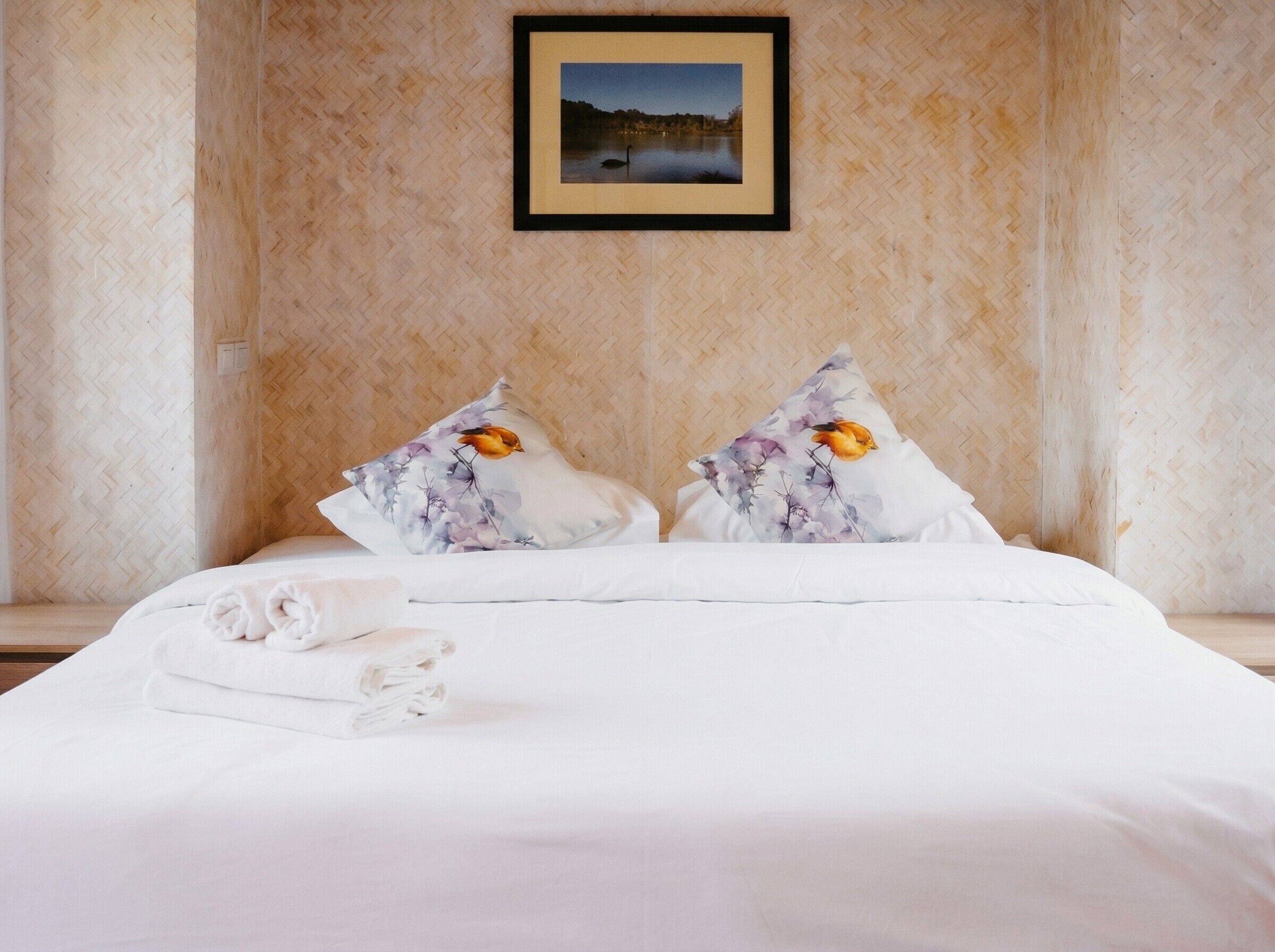 A neatly made bed with white linens, two pillows with floral designs, and a framed picture on the wall.