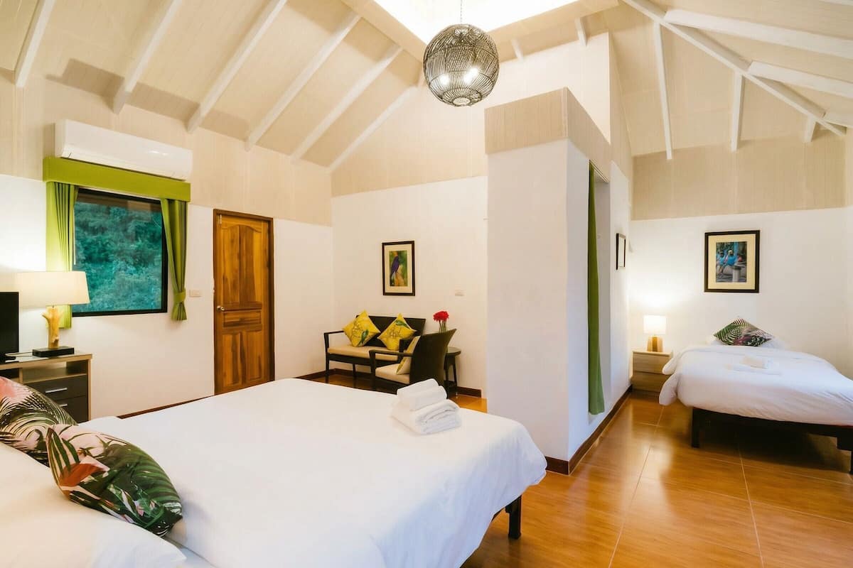 A hotel room with two beds, a wooden ceiling, a green curtain, a painting, and a window with a view.