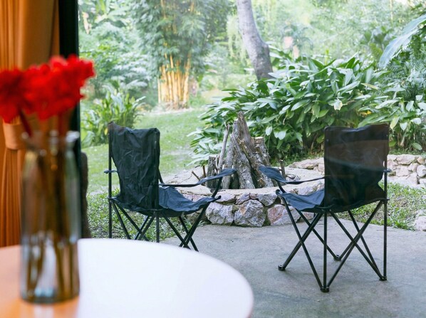 Un espacio al aire libre con dos sillas y una mesa, rodeado de follaje y un hogar de piedra.